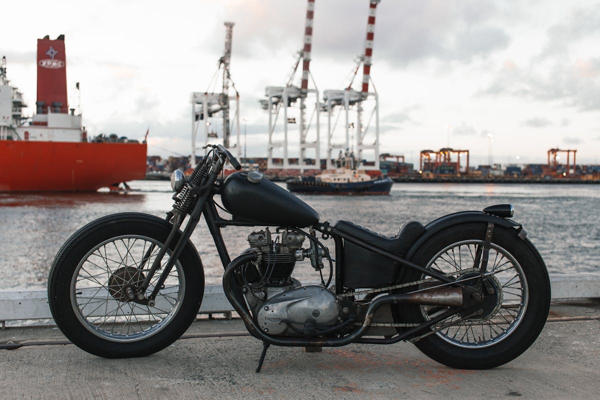 Classic motorcycle parked by a busy harbor with cranes and ships in the background.
