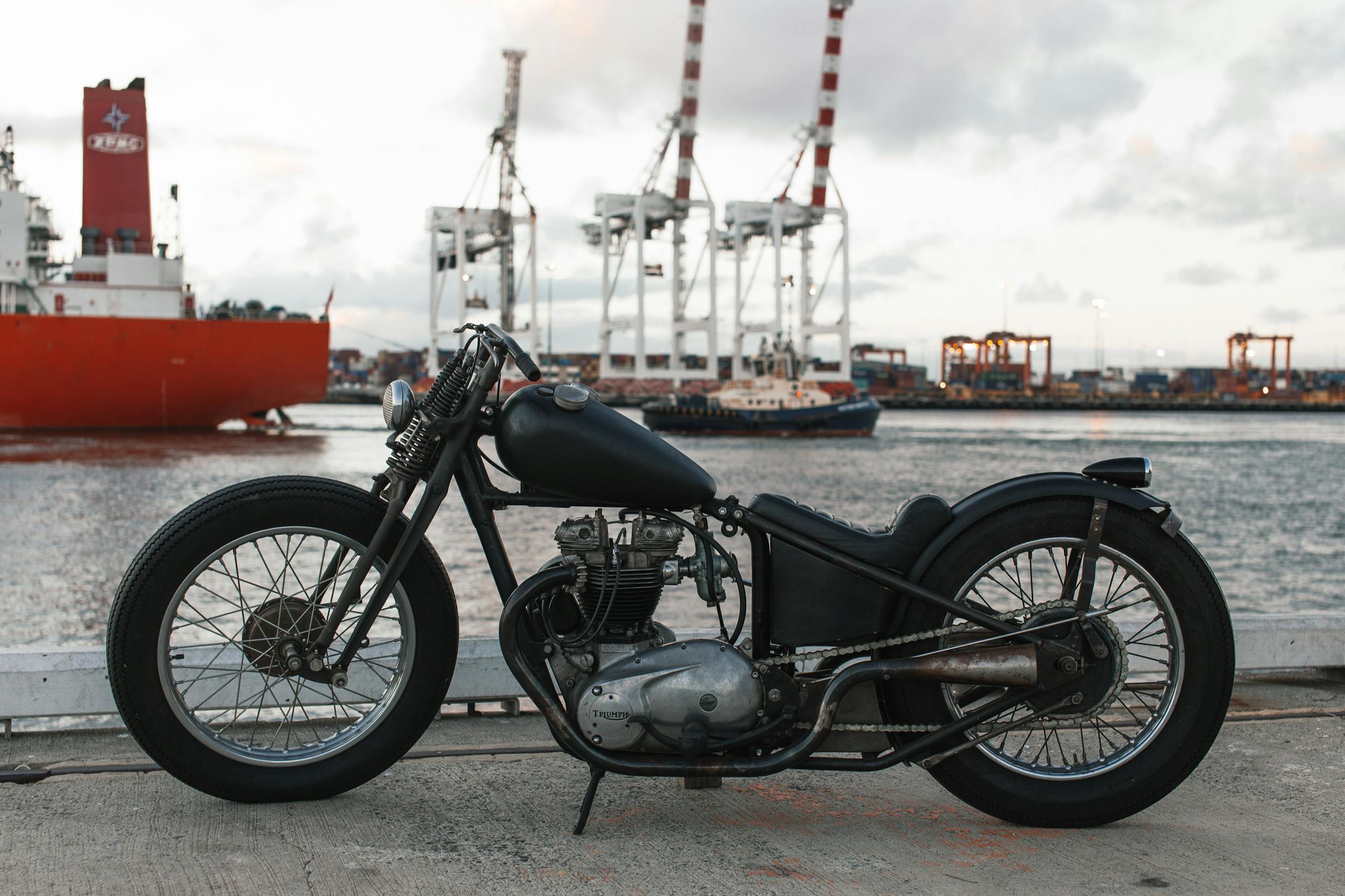 Classic motorcycle parked by a busy harbor with cranes and ships in the background.